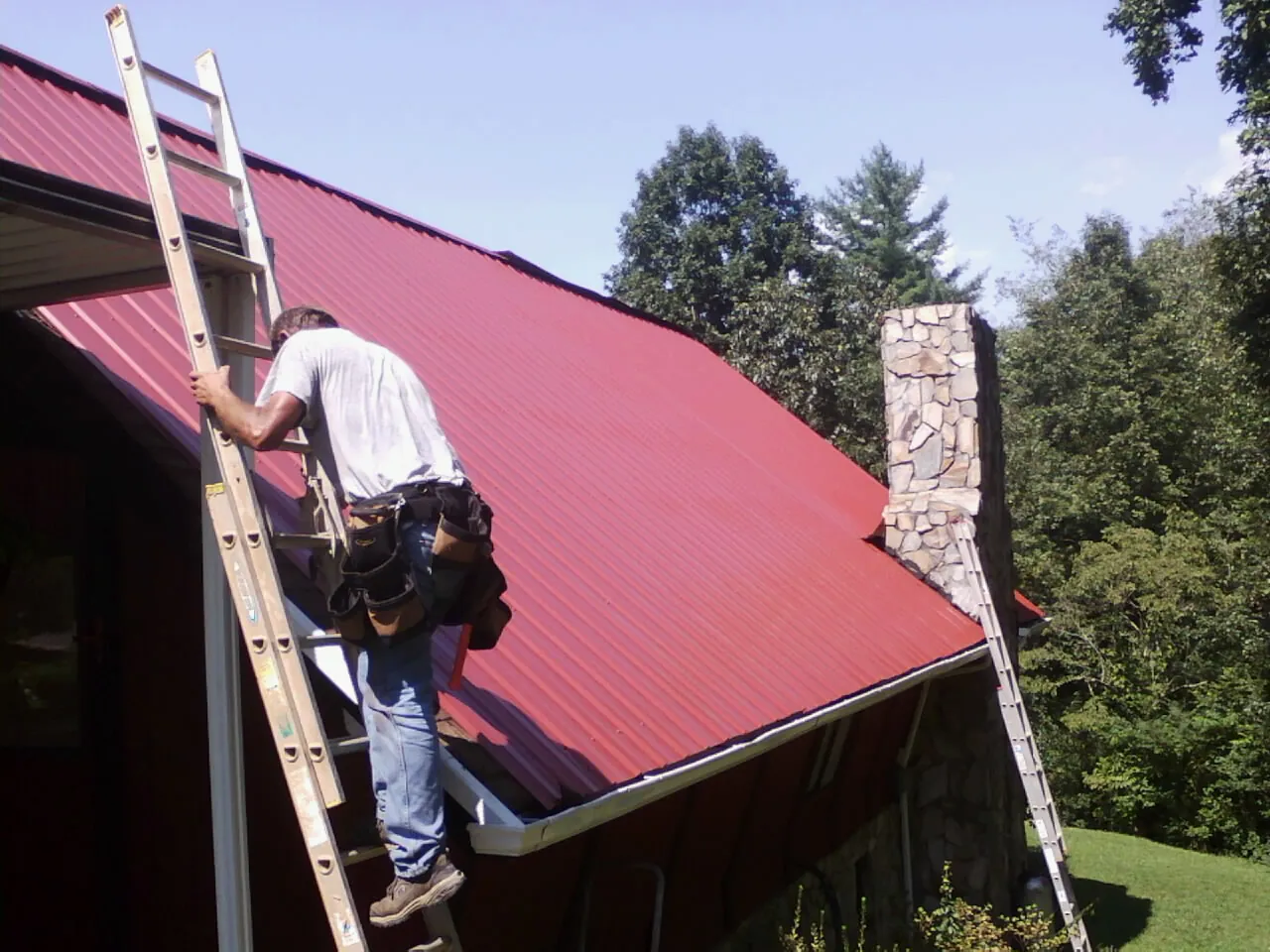 Roofer on a ladder beside a red metal roof and stone chimney — Soffit & Fascia Repair by Roofing Brothers in Palisades Park
